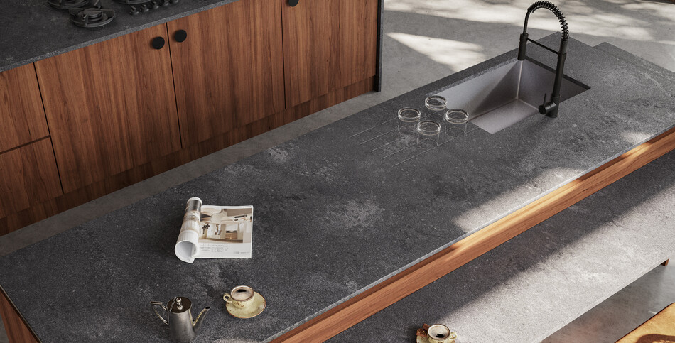Coffee cups and a magazine sit atop a Rugged Concrete quartz kitchen island of robust grays flushed with white-haze patinas and industrial-inspired imperfections.